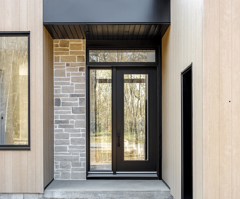 Porte d’entrée extérieure Vaillancourt Portes et Fenêtres de couleur noire avec verre clair latéral et imposte agencée à des revêtements de pierre et de bois blond